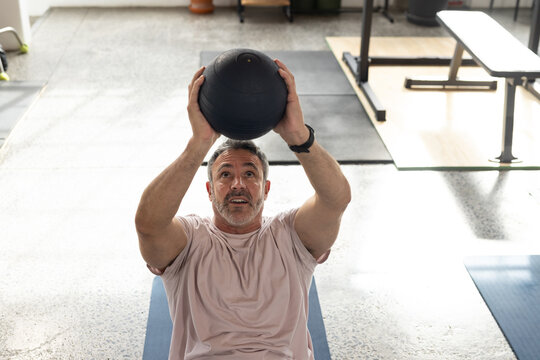 Middle-aged man sitting on blue exercise mat in gym holding black medicine ball overhead