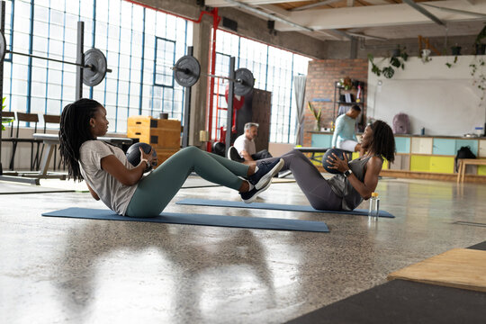 African American women exercising on blue mats in converted warehouse gym, holding medicine balls