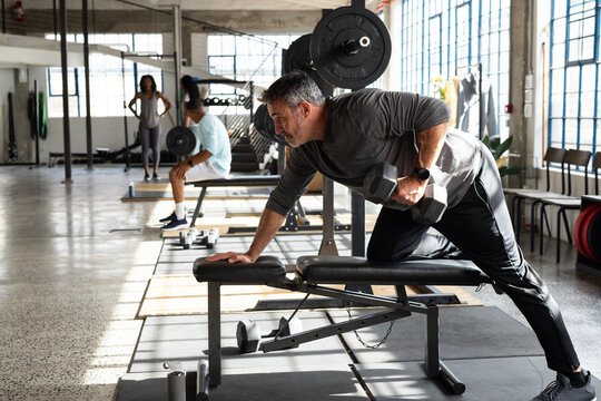 Mid adult man performing one-arm row with heavy hex dumbbell on adjustable bench in converted gym