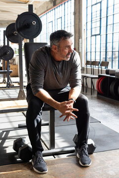Middle-aged man sitting on flat bench in gym near loaded barbell wearing long-sleeve top and watch