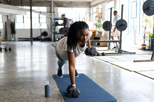 African American woman in teal leggings performing plank row on mat at gym with hex dumbbells