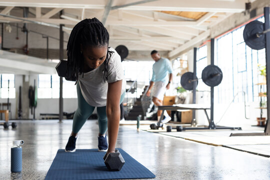 Diverse gym members working out in bright gym, woman performing single-arm dumbbell row on blue mat