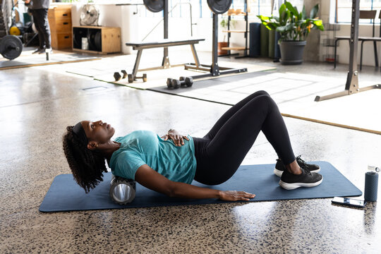 Mid-adult African American woman in teal-top rolling on roller on mat at gym bottle phone nearby