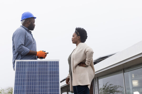 African American man and woman inspecting foreground solar panel on roof, man wearing blue hard hat