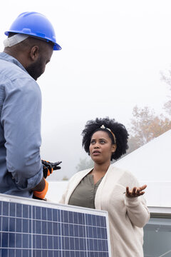 African American woman and man discussing roof solar panel, man wearing blue hard hat orange gloves