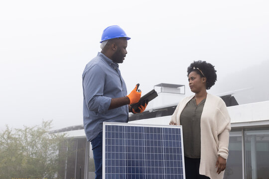Man tech and woman client discussing solar panel on roof with tablet, man wearing hard hat