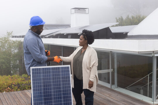 African technician wearing orange gloves and homeowner shaking hands on roof beside portable panel