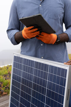 Diverse technicians inspecting solar panel on rooftop, using tablet and wearing orange gloves