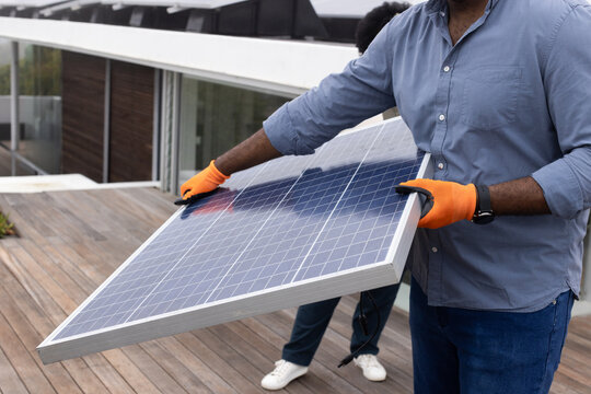 African American coworkers lifting and positioning framed solar panel on rooftop deck with gloves