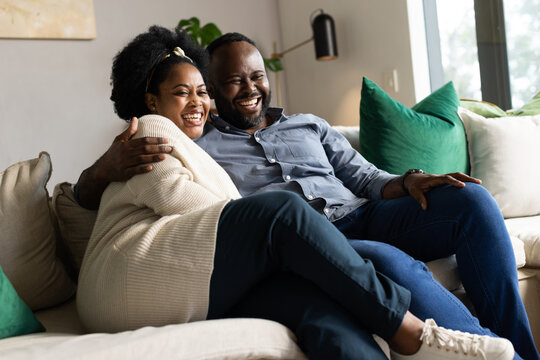 African couple sitting on beige sofa leaning together laughing by large window with green pillows