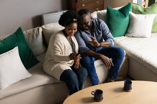 African couple sitting on light-colored sectional sofa in living room holding smartphone, smiling