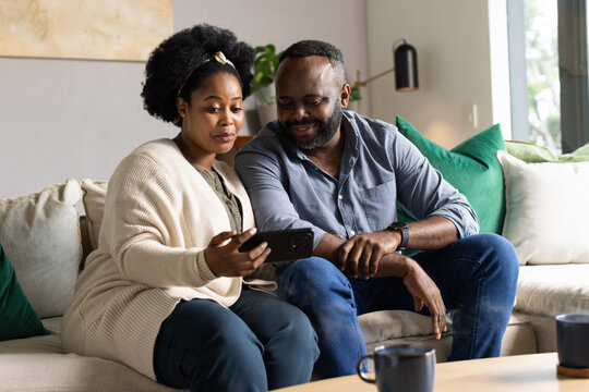 African American couple sharing smartphone screen while sitting on beige sofa in living room