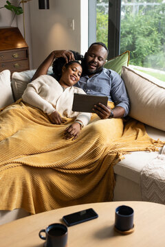 African American couple reclining on sofa at home under mustard-yellow throw, man holding tablet