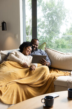 African American couple sitting on sectional sofa, sharing tablet under mustard knit blanket