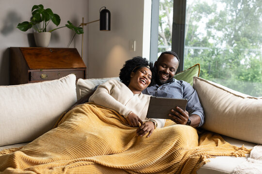 African couple reclining on light sectional sofa, smiling and holding tablet under mustard throw