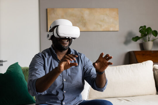 African adult man sitting in living room wearing blue shirt, VR headset, gesturing at virtual items