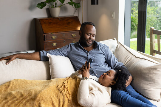 African American couple cuddling on light-colored sofa in living room, woman scrolling smartphone
