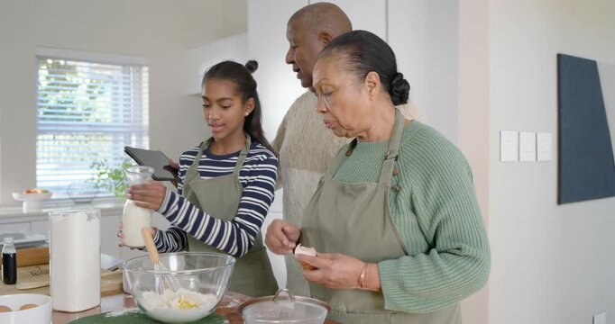 African American seniors guiding teen mixing batter at kitchen island, checking tablet, adding milk