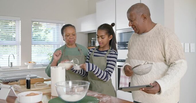Grandparents guiding grandchild following tablet, measuring sifting mixing flour at kitchen island