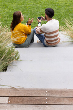 African American friends sitting on wide steps on lawn holding dark mugs, wearing mustard sweater