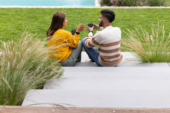 African American couple sitting on concrete steps by pool, wearing sweaters and holding dark mugs