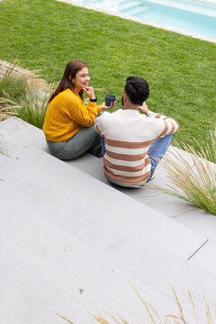 African American couple wearing mustard and striped sweaters holding mugs on concrete steps by pool