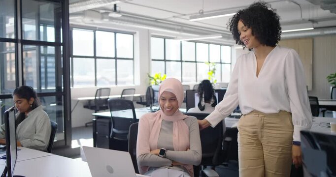 Diverse female team in office, peer gesturing at laptop, pink hijab woman showing typing for review