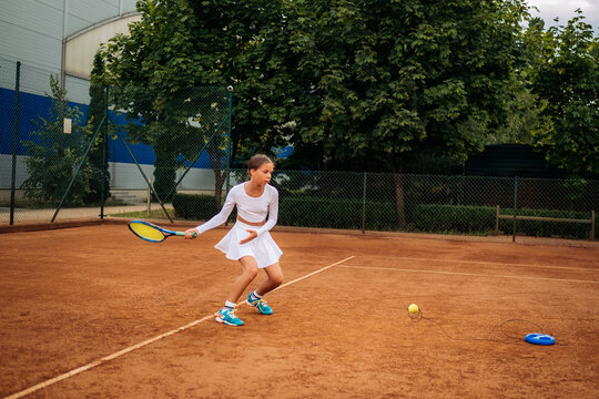 Young girl playing tennis on an outdoor clay court with a racket, practicing action shot in white activewear outfit.