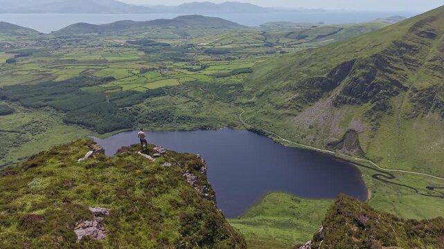 Cinematic aerial view of drone flying past a man standing on a cliff edge revealing Annascaul Lake on Dingle Peninsula County Kerry Ireland