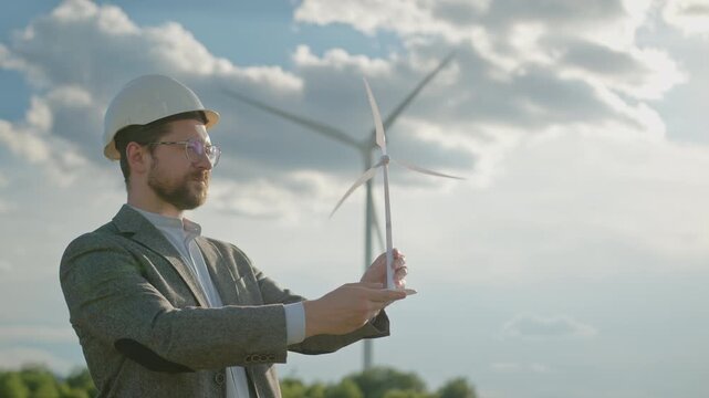 Man in hard hat and suit holds miniature wind turbine, examining it with wind turbines in background under cloudy sky. Engineer analyzing sustainable technology project