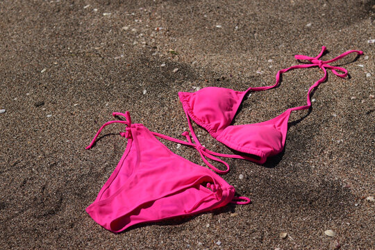 Bright pink women's one-piece swimsuit bikini lies on a sandy beach under the sun's rays. Pink triangle swimsuit with ties, spread out on dark sea sand. Summer vacation concept