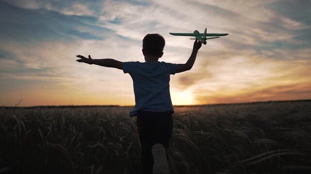Silhouette of happy boy running through field with green toy airplane at sunset. Child holds plane raised high with arm outstretched. Kid runs across grass toward sunset sky, dreaming of flight.