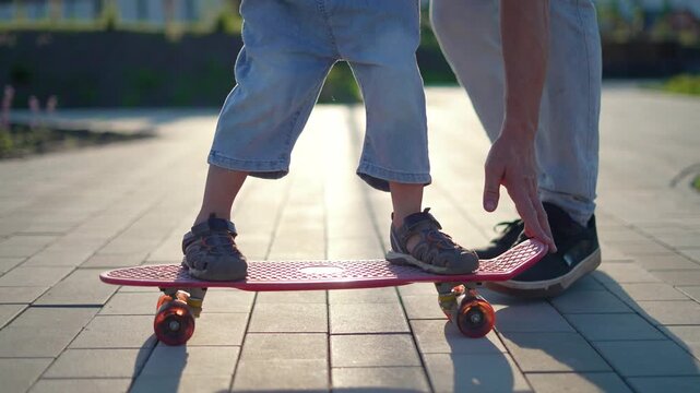 Father teaching child skateboarding on paved sidewalk at sunset. Curious child learns to balance on pink skateboard. Parent hand holds board steady. Family skateboarding lesson outdoor in park.
