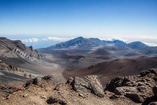 Vast volcanic crater stretches under bright blue sky. Rugged rocks frame foreground with sharp, weathered texture. Distant mountains loom beyond misty cloud layers. Sunlight bathes the terrain in warm
