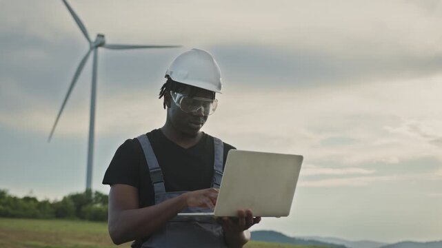 African American engineer wearing hard hat and safety glasses checks data on laptop near wind turbines in field. Footage highlights renewable energy monitoring and technical analysis