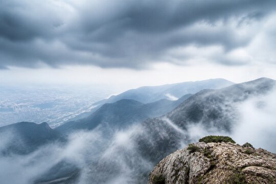 Stormy clouds loom over mist-shrouded mountains. Rocky peak juts from fog, crowned with sparse green life. Distant city sprawls beneath rolling hills. Mist curls between peaks, soft and silent