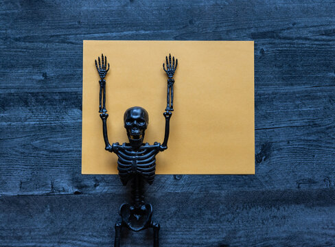 Funny halloween concept flat lay background blank empty yellow sheet of paper with black plastic skeleton with arms raised. 
