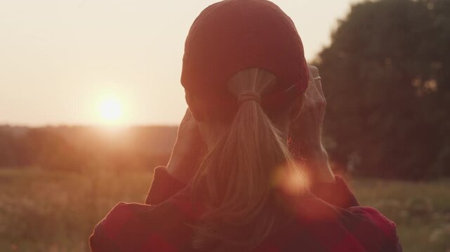 Young woman in a red cap watches the sunset over a rural landscape, viewed from behind.
