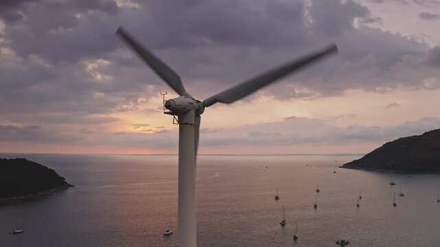 Wind turbine generating clean energy power, blades rotating at colorful sunset over the Andaman Sea with sailboats docked near the mountain coast of Phuket, Thailand. Aerial drone view static shot