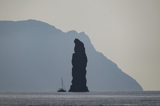 Silhouette of a towering rock needle, behind it the island of Alicudi, a sailboat in the calm sea near Filicudi, Aeolian or Lipari Islands, southern Italy, Italy