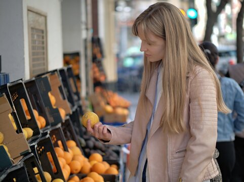 Young woman in a beige leather jacket picks oranges from crates at a fruit stall on the pavement, a sunny street with passers by behind her