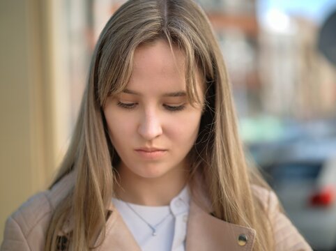 Young woman with long blonde hair lowers her eyes in thought on a sunny street, a pale yellow wall and blurred city behind her