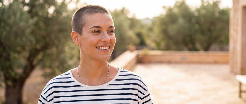 Smiling woman with buzz cut on terrace enjoying warm golden hour sunlight in a striped t-shirt, perfect for travel, modern lifestyle, wellbeing, and body positivity campaigns