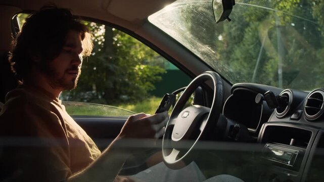 Man checks his smartphone while sitting behind the steering wheel in warm evening light. Automobile interior and mobile communication suggest an insurance inquiry or customer service call.