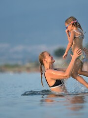 Fototapeta premium Woman in a black swimsuit lifts her small daughter high above the calm sea, the child laughing with her arms out against the sky