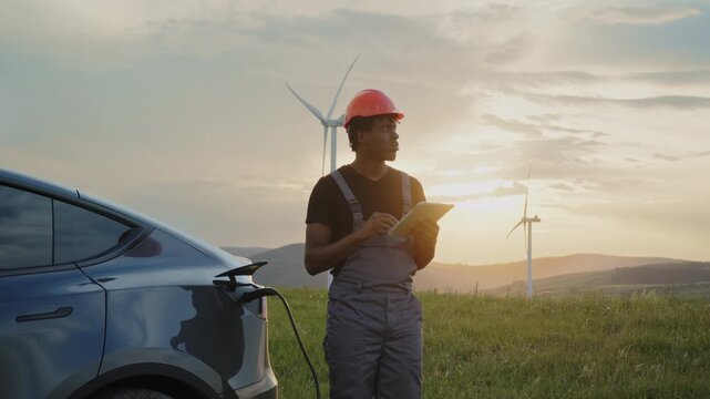 Man in hard hat and overalls checks tablet near electric car charging at wind farm during sunset. Footage illustrates renewable energy and eco-friendly transport