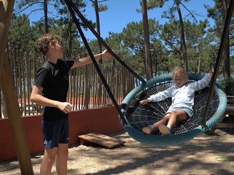 Boy pushes a round rope nest swing while a girl sits inside, sunny pine forest playground