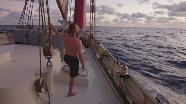 Sailing the Atlantic. Male sailor stands on the deck of the big sailing boat moving in the Atlantic Ocean under the sails and enjoys the first light of the day