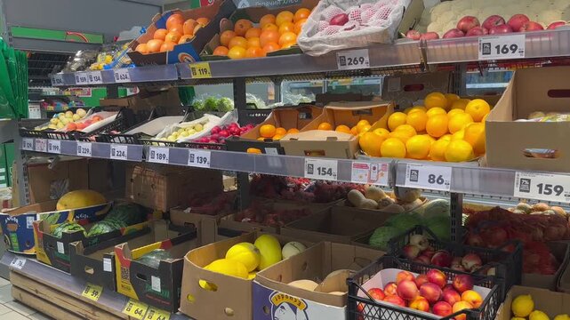 Bright oranges in cardboard boxes line supermarket shelves. Price tags show varying costs across different produce sections. Fruit displays feature apples, tomatoes, and leafy greens