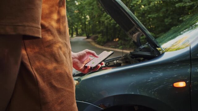 Man uses a smartphone beside a disabled vehicle in the forest, contacting an insurer or emergency support after an accident or mechanical failure. Roadside assistance and quick communication during